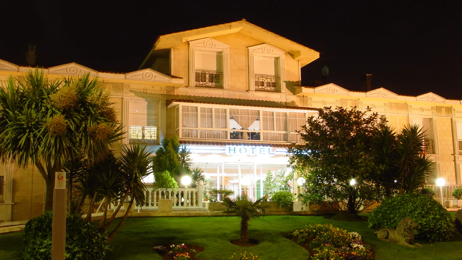 Fachada iluminada de un hotel cl&aacute;sico, rodeado de plantas y jardines, de noche.