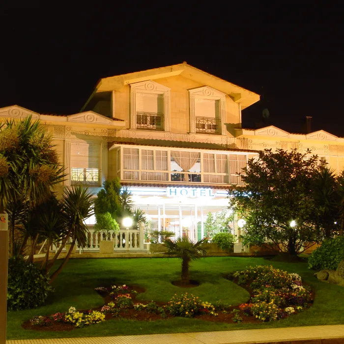 Hotel iluminado de noche con jard&iacute;n frontal y varias plantas.