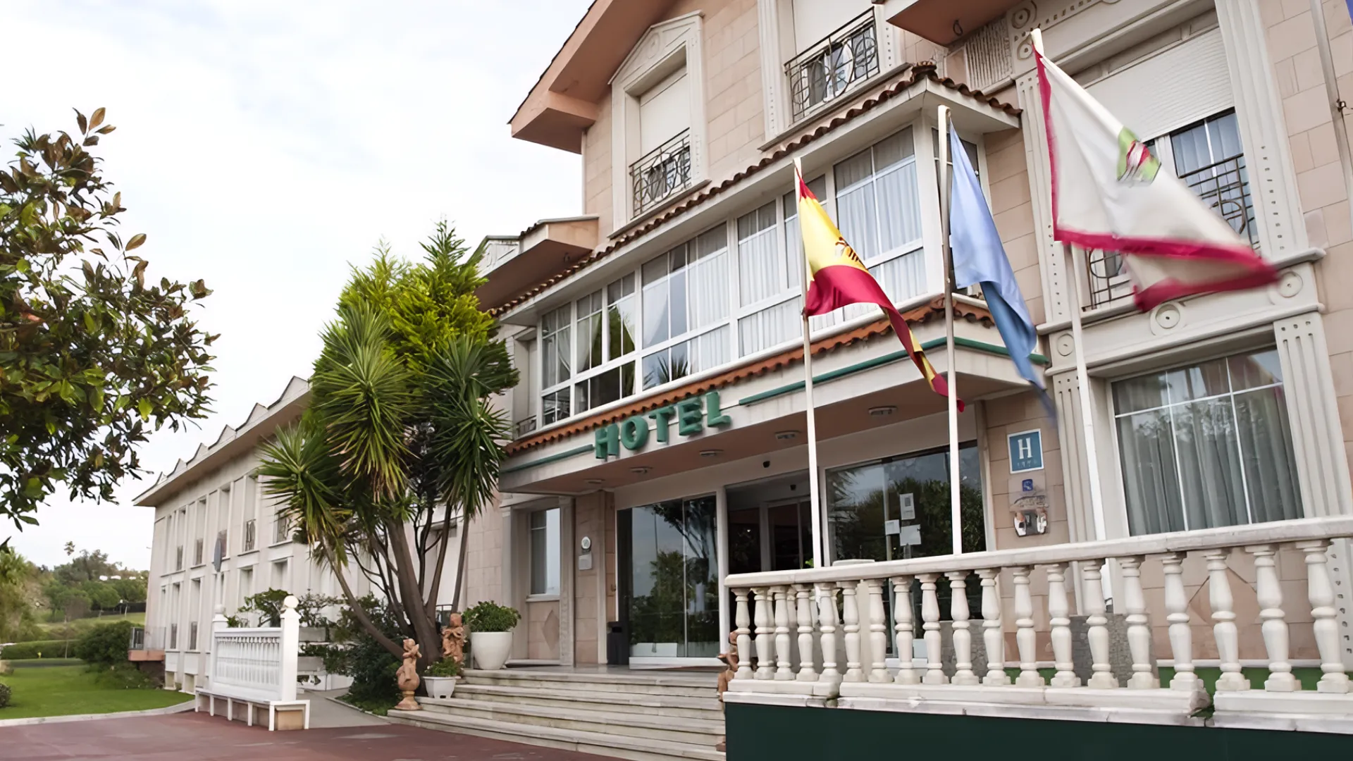 Fachada de un hotel con jard&iacute;n y banderas ondeando.