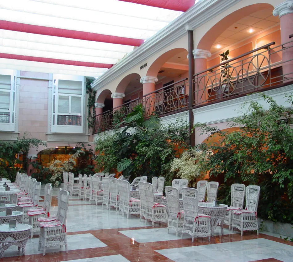 Patio interior con sillas y mesas blancas de mimbre, rodeado de plantas y con techo acristalado.