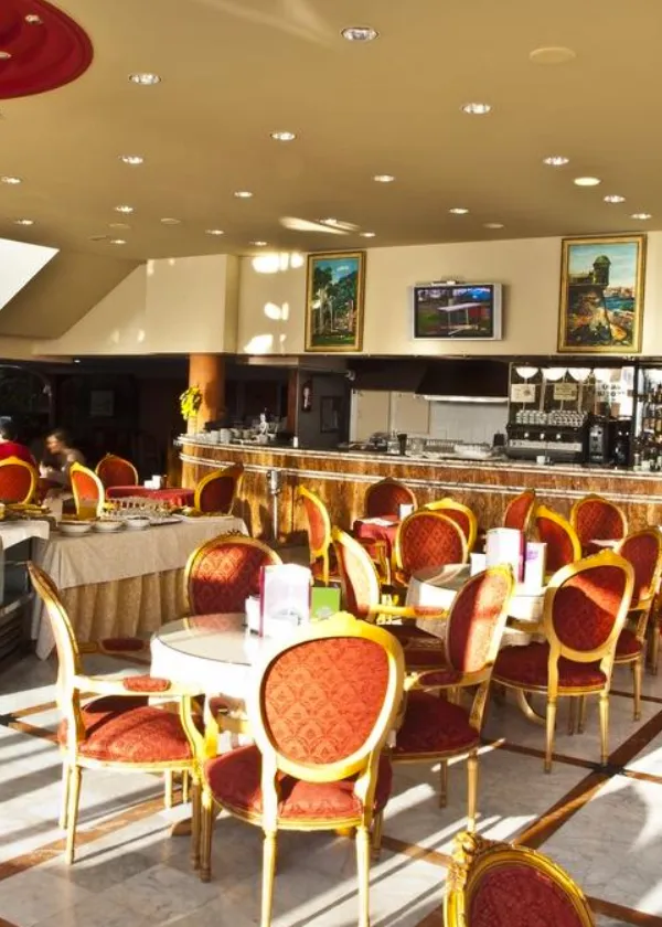 Cafeter&iacute;a elegante con sillas rojas y mesas redondas, barra al fondo y decoraci&oacute;n cl&aacute;sica.