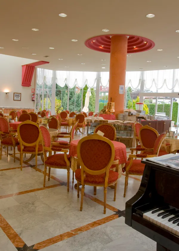 Restaurante elegante con sillas rojas, mesas redondas, piano y grandes ventanales.