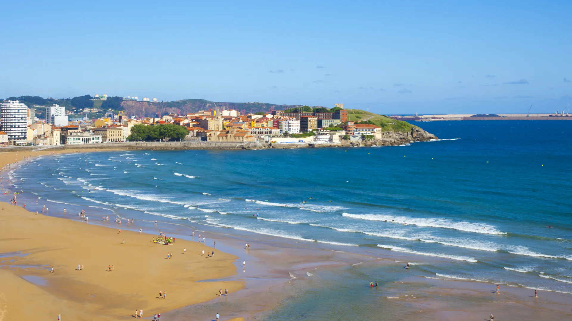 Playa con olas suaves y gente caminando, bordeada por una ciudad costera bajo un cielo despejado.
