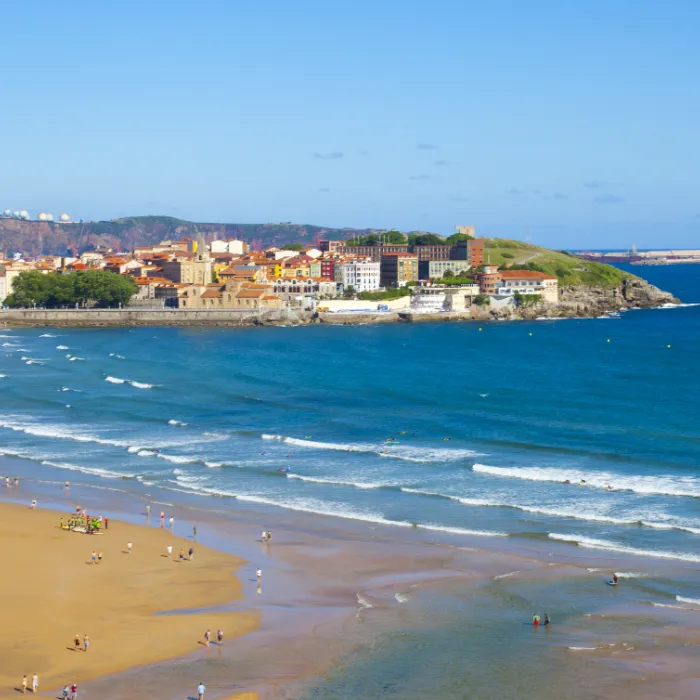 Vista de una playa con personas y una ciudad costera al fondo, con cielo azul y olas suaves.