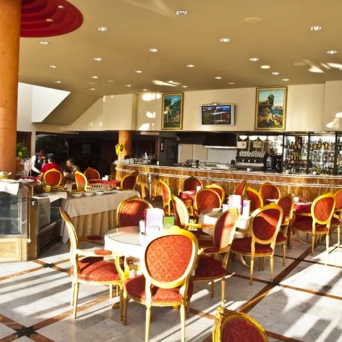 Cafeter&iacute;a elegante con sillas rojas, mesas redondas y un bar al fondo iluminado por luz natural.