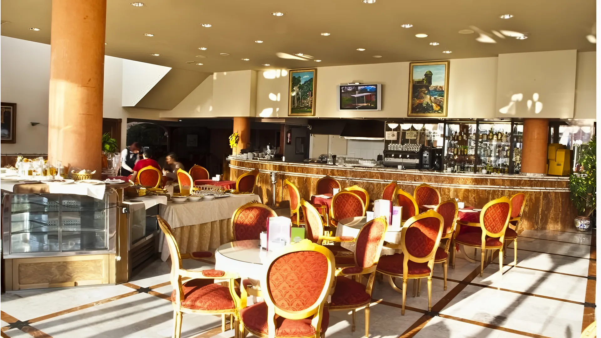 Cafeter&iacute;a elegante con sillas rojas, mesas redondas, barra, cuadros y techo con luces.