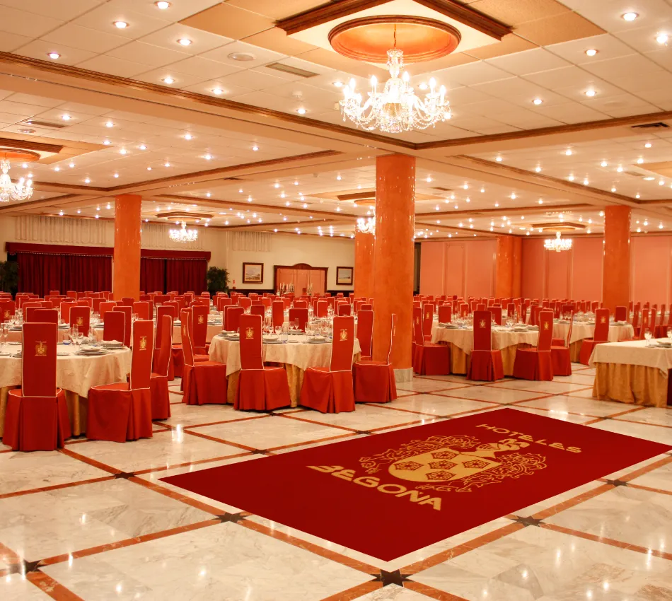 Gran sal&oacute;n de eventos con mesas y sillas rojas, iluminaci&oacute;n elegante y alfombra con logotipo.