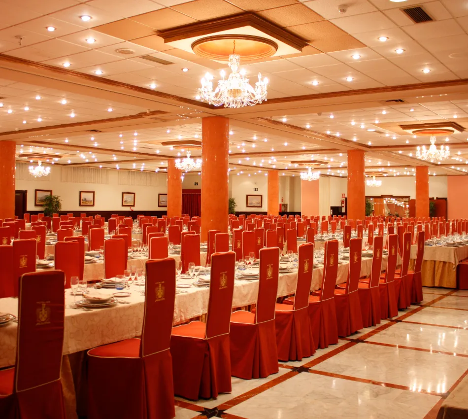 Sal&oacute;n de banquetes elegante con mesas y sillas rojas bajo candelabros brillantes.