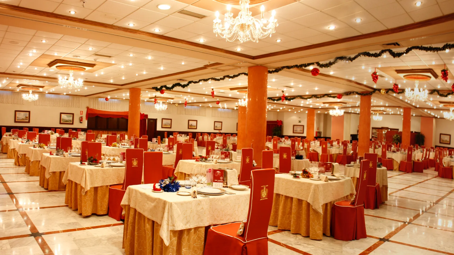 Sal&oacute;n de banquetes decorado con iluminaci&oacute;n c&aacute;lida, mesas arregladas y sillas con fundas rojas.