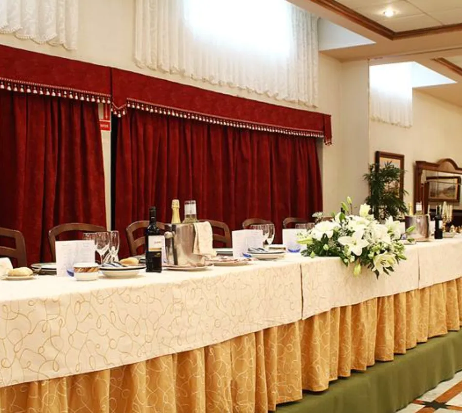 Mesa larga decorada con flores, copas y botellas. Cortinas rojas de fondo en un sal&oacute;n elegante.
