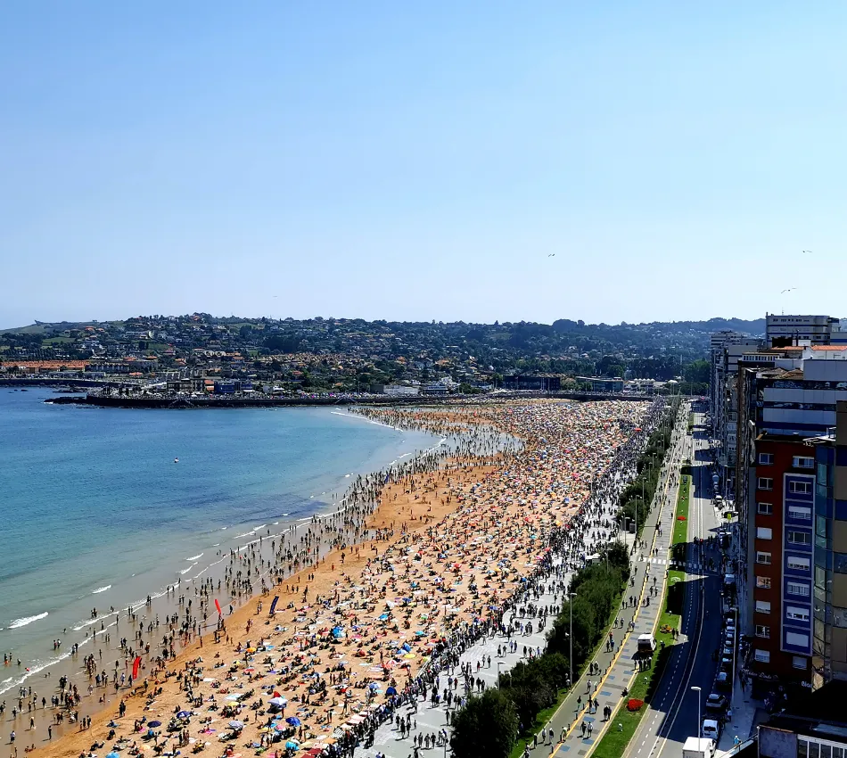 Playa abarrotada con sombrillas y personas junto a la ciudad y el mar bajo un cielo despejado.