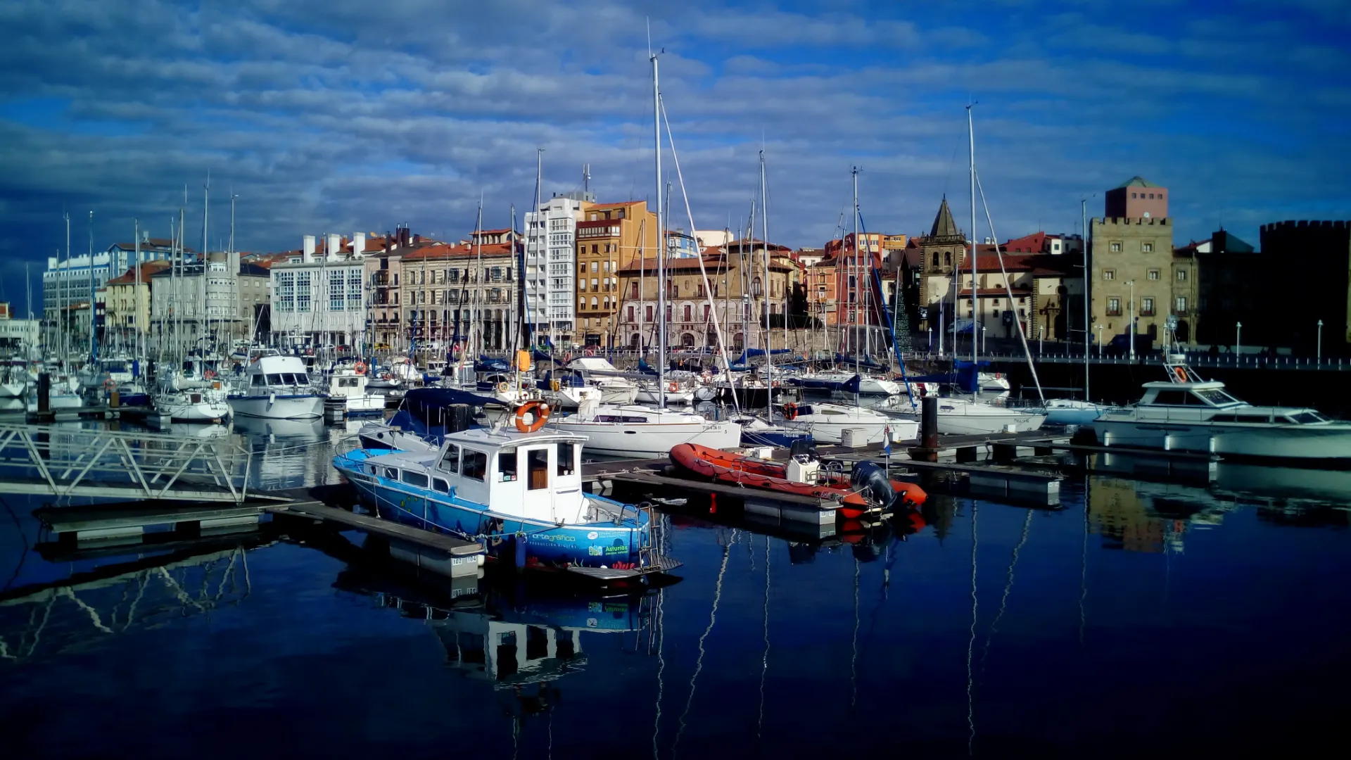 Puerto con barcos atracados y edificios al fondo bajo un cielo parcialmente nublado.