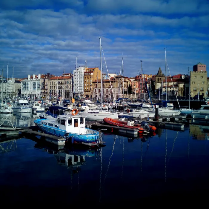 Puerto con barcos y edificios al fondo bajo cielo azul con nubes.