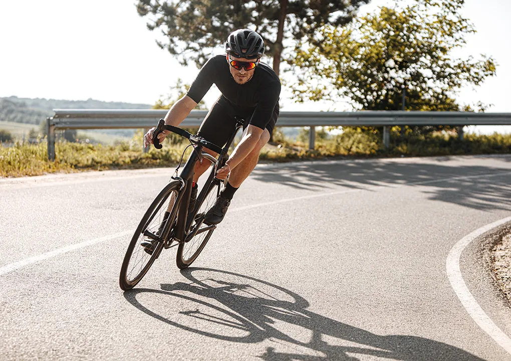 Ciclista con casco y gafas en una carretera curvada bajo el sol.