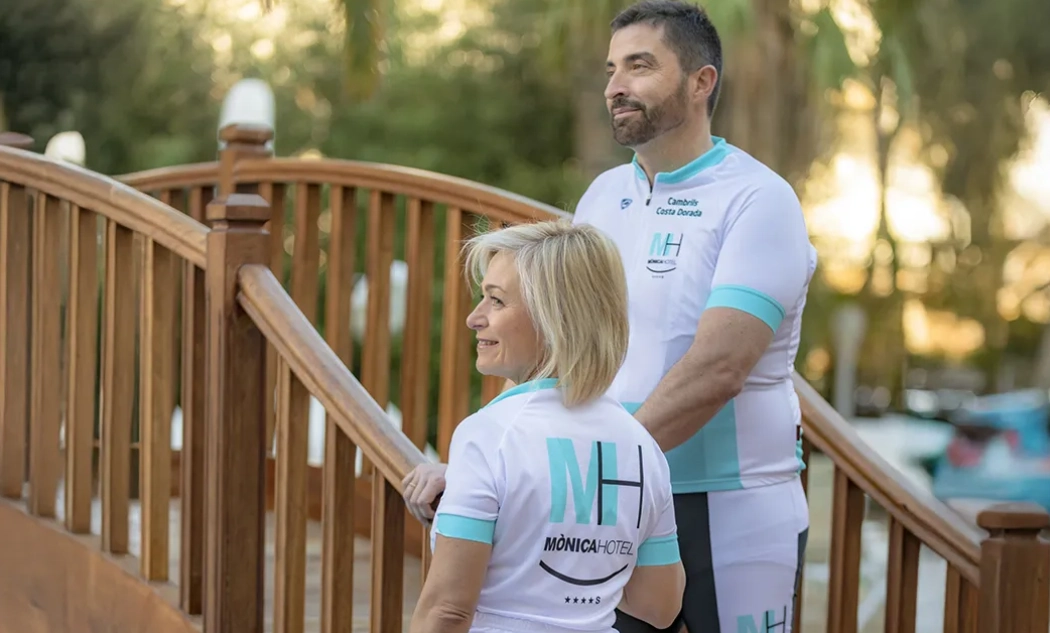 Dos ciclistas en un puente de madera, visten uniformes de M&oacute;nica Hotel.