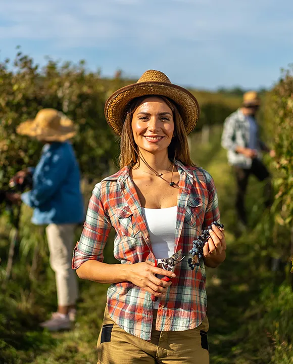 Mujer sonriendo en un vi&ntilde;edo con uvas en la mano y sombrero de paja.