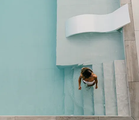 Mujer descendiendo escaleras de una piscina con silla blanca en el borde.