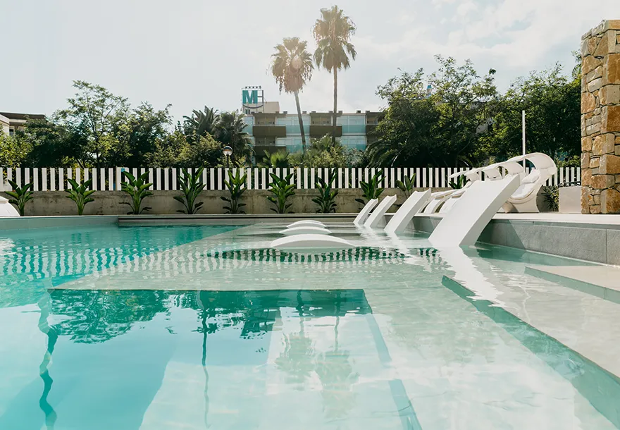 Piscina con reposeras sumergidas y palmeras al fondo en un entorno soleado.