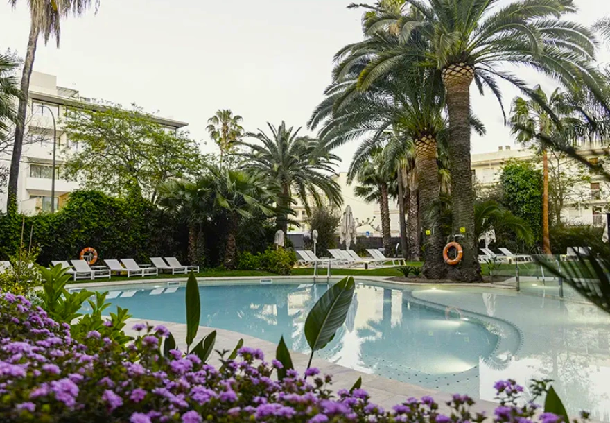 Piscina rodeada de palmeras y flores, con tumbonas blancas alrededor.