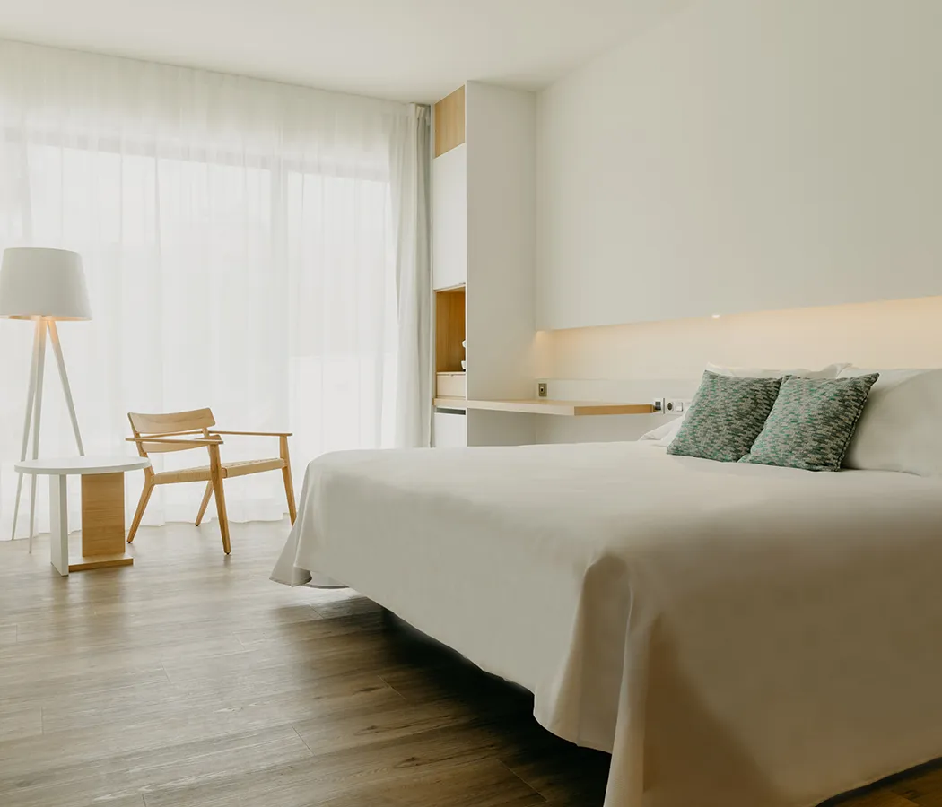 Habitaci&oacute;n minimalista con cama, silla, l&aacute;mpara y cortinas blancas.