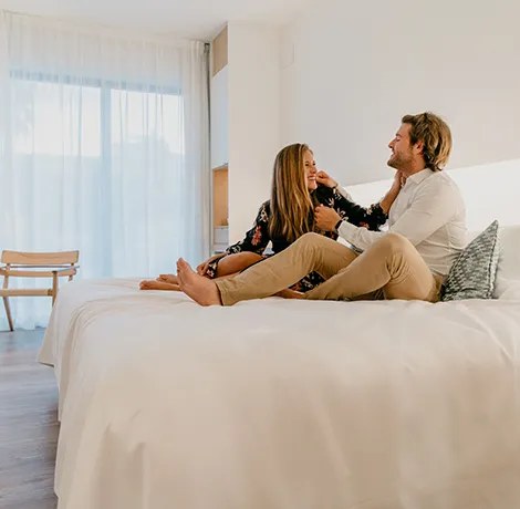 Pareja sonriendo sentada en una cama en una habitaci&oacute;n luminosa.