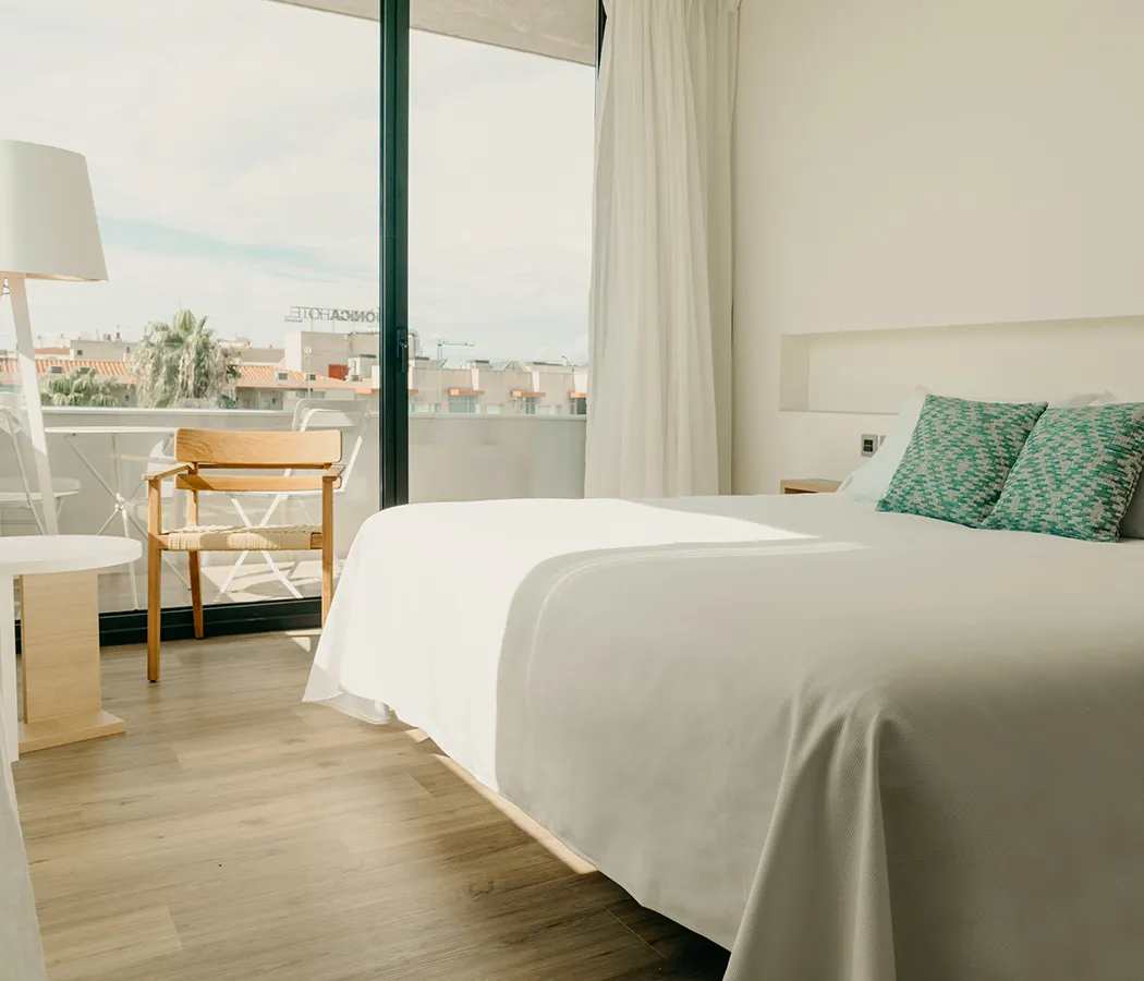 Habitaci&oacute;n luminosa con cama, almohadas verdes y ventana al balc&oacute;n.