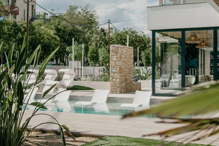 Piscina con tumbonas blancas rodeadas de vegetaci&oacute;n y paredes de vidrio.
