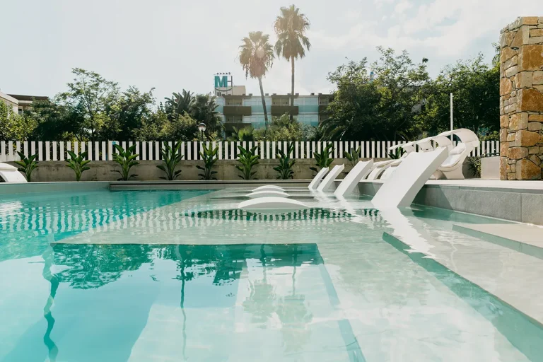 Piscina moderna con sillas de dise&ntilde;o bajo palmeras al fondo.