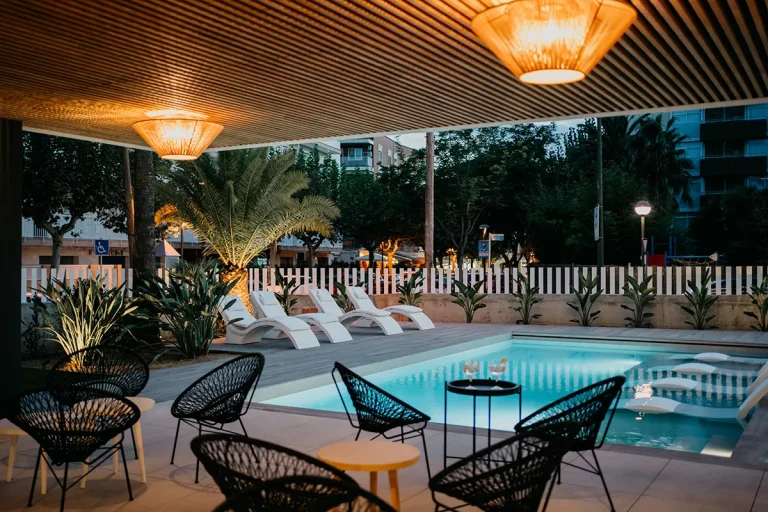 Piscina iluminada al anochecer con sillas y plantas alrededor.