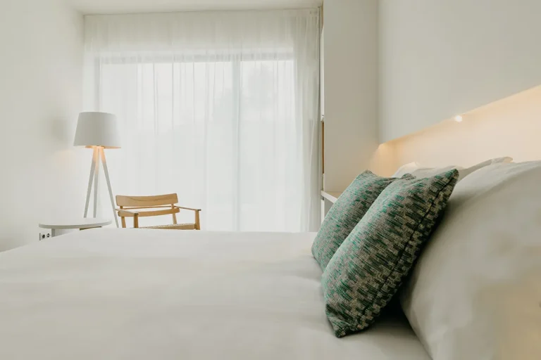 Dormitorio minimalista con cama, cojines, l&aacute;mpara y silla junto a ventana.