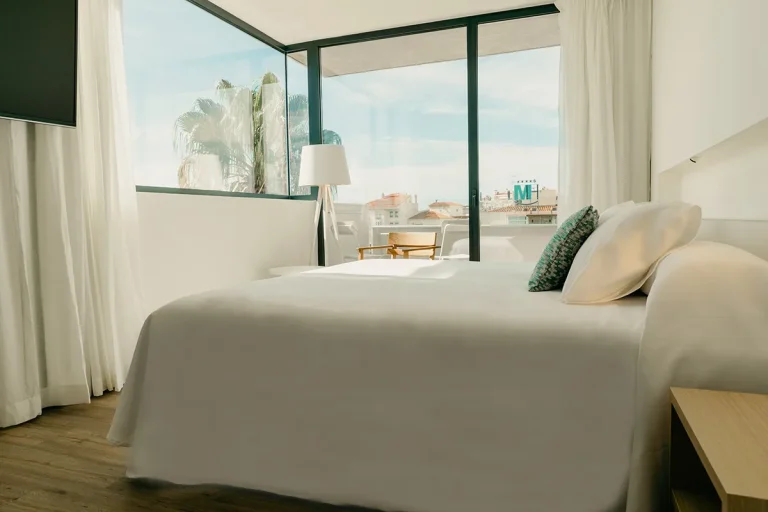 Habitaci&oacute;n luminosa con cama blanca y ventana panor&aacute;mica.