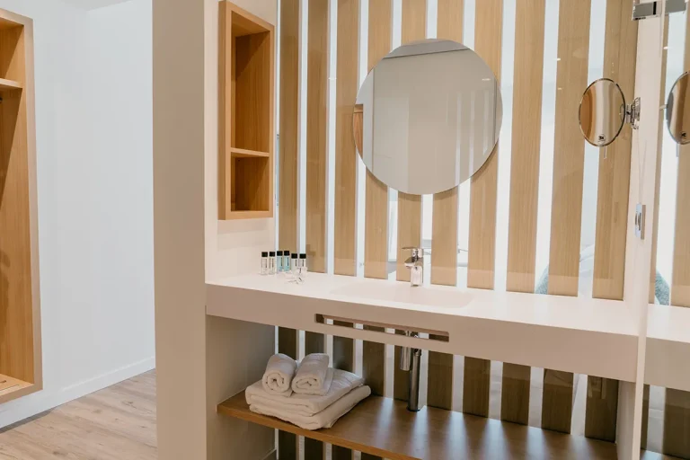 Ba&ntilde;o moderno con espejo circular y toallas blancas en un estante de madera.