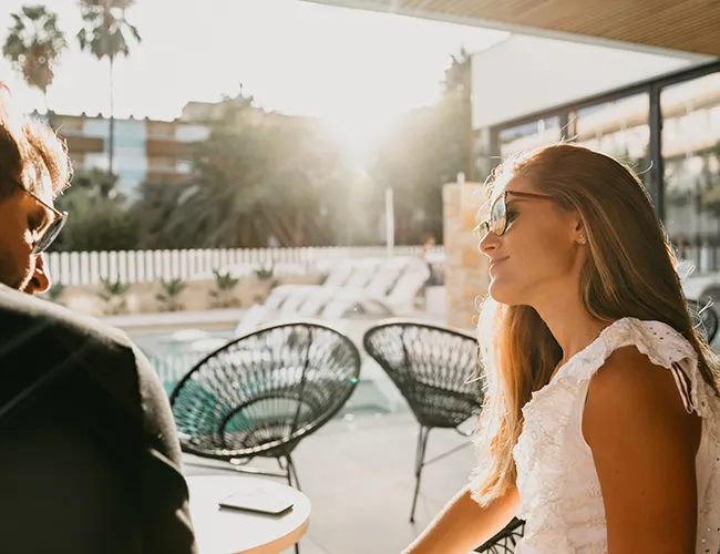 Mujer con gafas de sol sonr&iacute;e en una terraza soleada junto a una piscina.