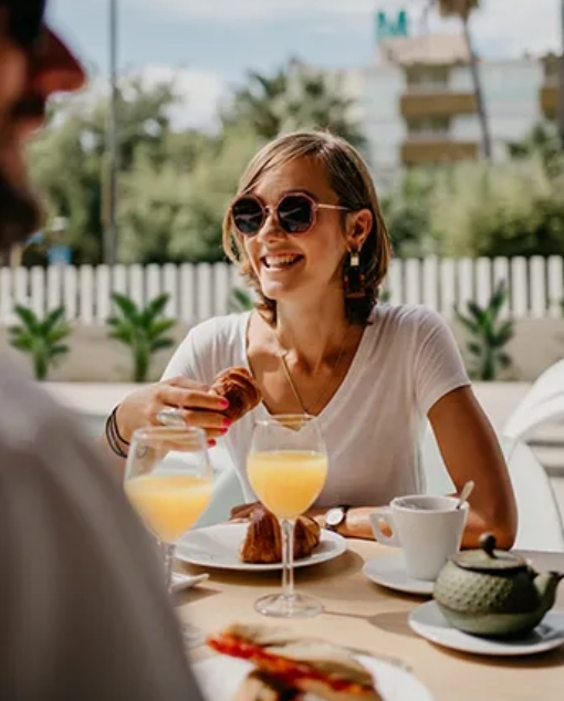 Persona sonriendo mientras desayuna al aire libre con jugo y croissant.