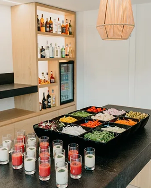 Mesa con ensaladas y bebidas, estanter&iacute;a con botellas al fondo.
