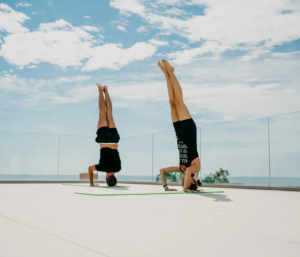 Dos personas haciendo yoga en una terraza junto al mar, en posici&oacute;n de invertida.