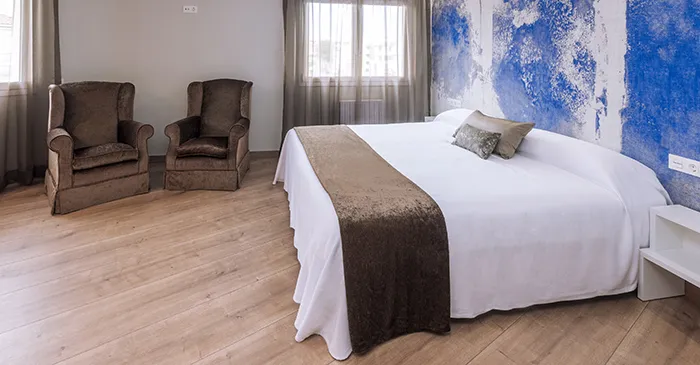 Habitaci&oacute;n con cama grande, sillas marrones y pared azul texturizada.