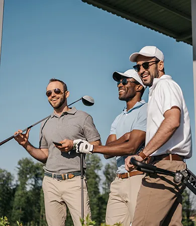 Tres hombres sonr&iacute;en con palos de golf al aire libre bajo un cielo azul.