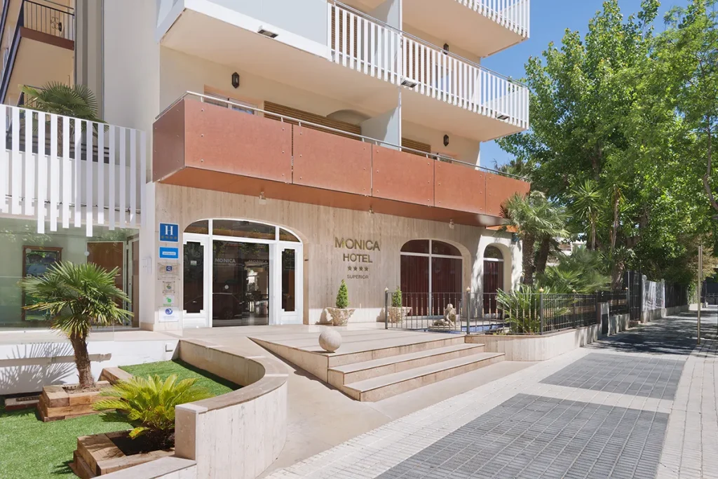 Entrada al Hotel con plantas y escalones, edificio moderno y fachada clara.