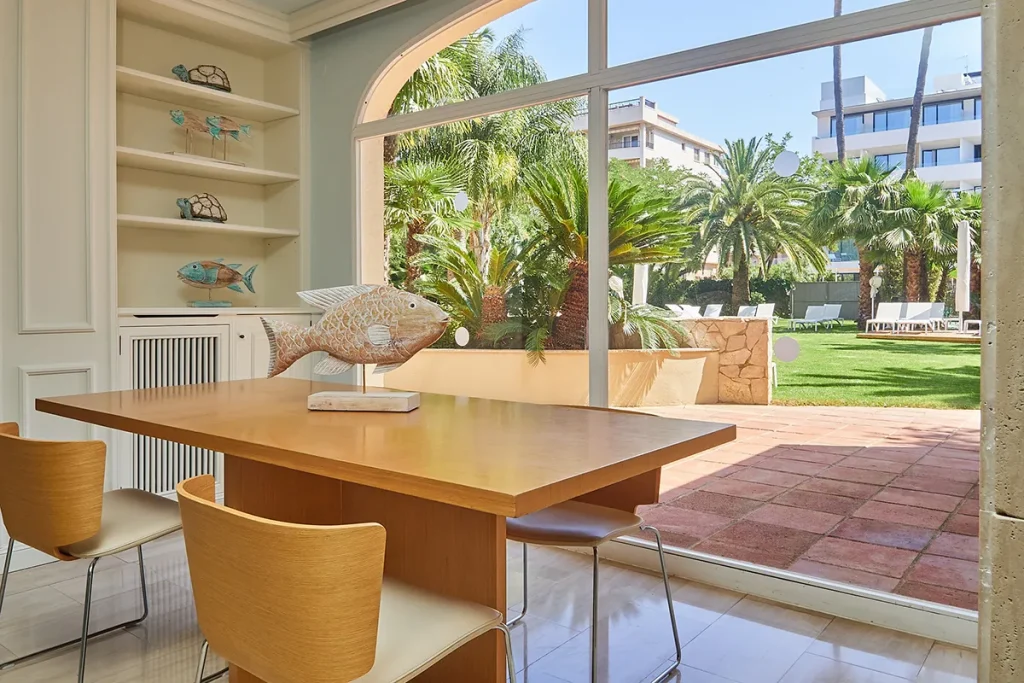 Sala con mesa, sillas y esculturas de peces, vista al jard&iacute;n con palmeras.