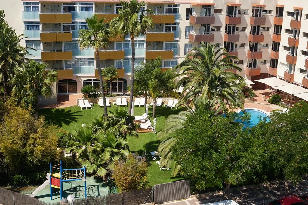 Hotel con jard&iacute;n, piscina, palmeras y &aacute;rea de juegos.