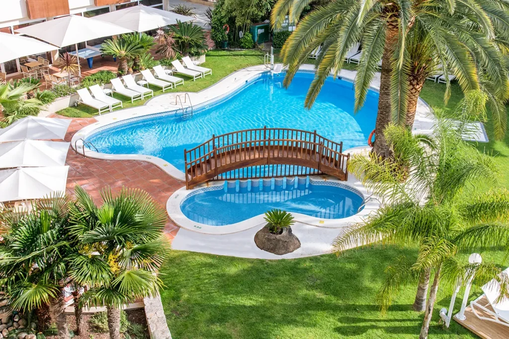 Piscina con puente de madera rodeada de palmeras y tumbonas blancas.