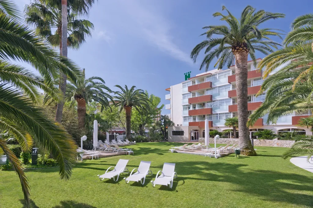 Jard&iacute;n de hotel con palmeras y tumbonas bajo un cielo despejado.