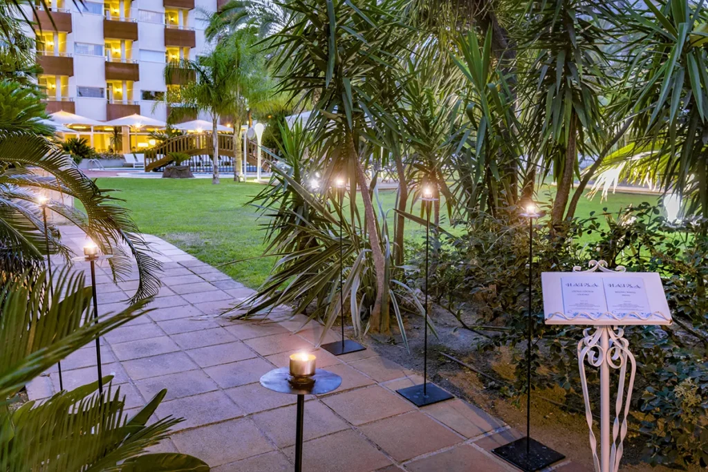 Camino iluminado por velas en un jard&iacute;n con plantas y edificio al fondo.