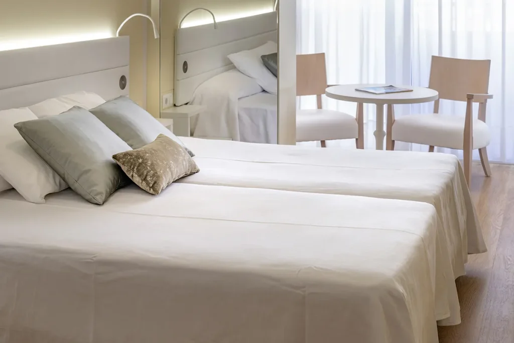 Cama doble con cojines en habitaci&oacute;n iluminada, mesas y sillas al fondo.