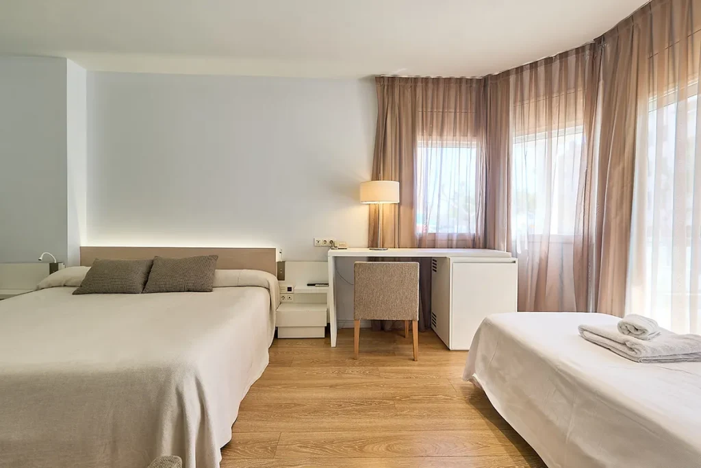 Habitaci&oacute;n moderna con dos camas, escritorio y l&aacute;mpara junto a una ventana.