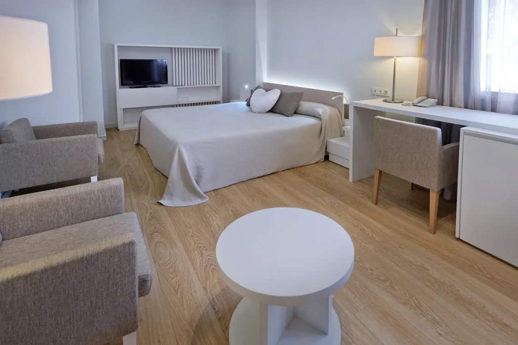 Habitaci&oacute;n moderna con cama, TV, escritorio y sillas grises.