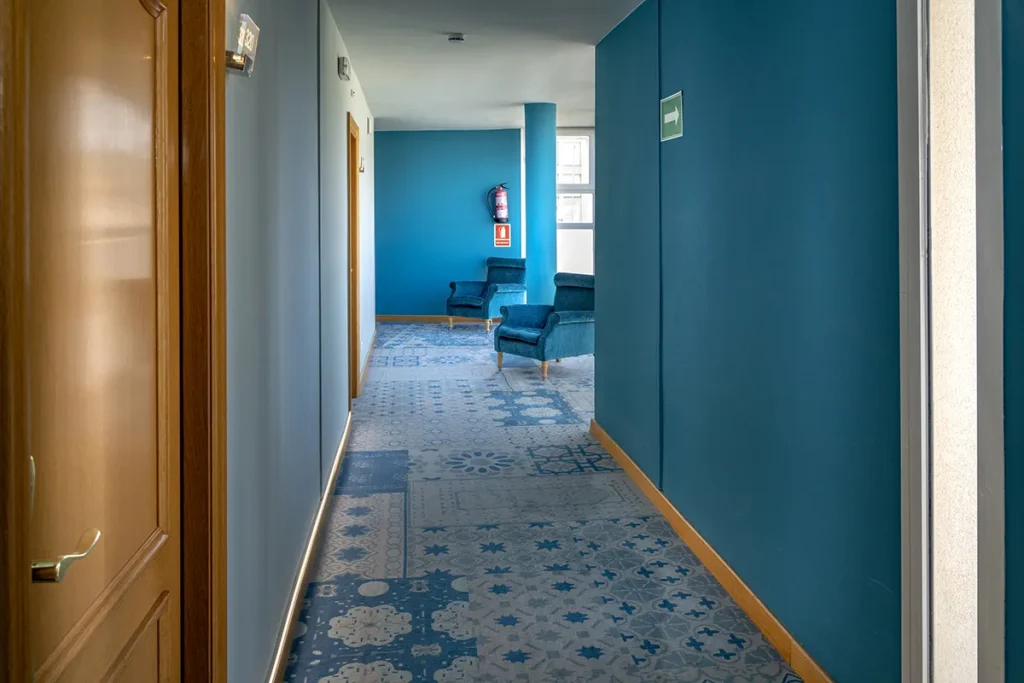 Pasillo azul con sillones al fondo y extintor en la pared.