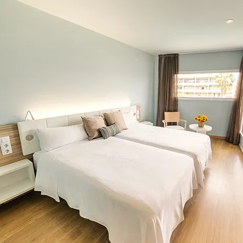 Habitaci&oacute;n luminosa con dos camas, mesa con flores y ventana con vista.