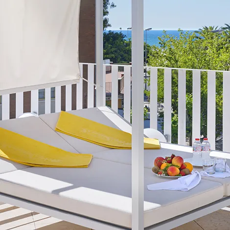 Cama al aire libre con toallas amarillas, frutas y vista al mar.